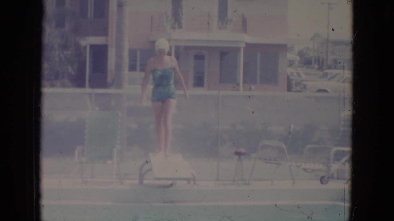 Woman in a teal swimsuit and white swim cap stands on a diving board by a pool.