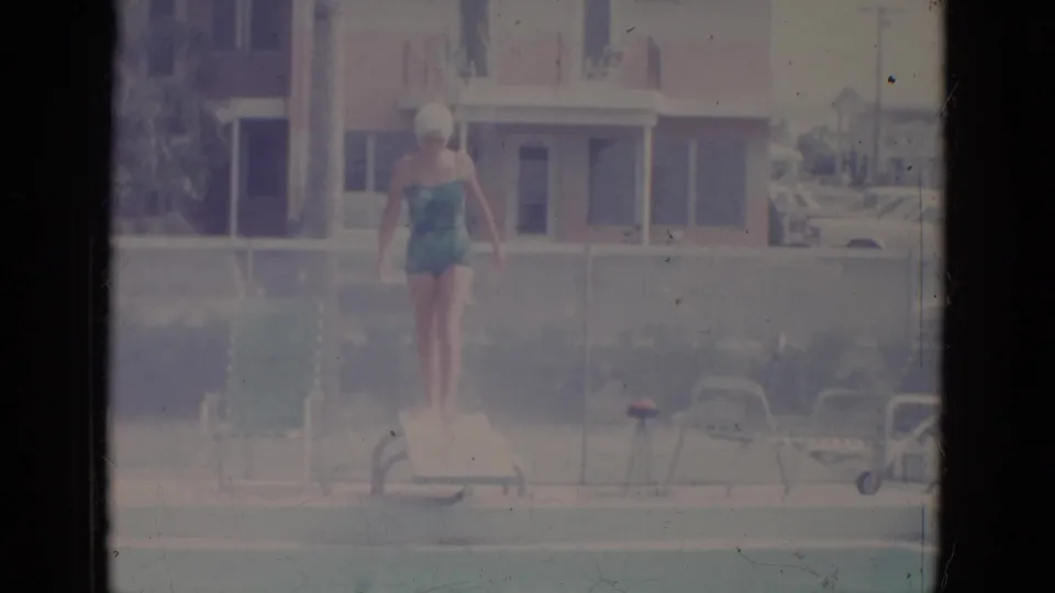 1966 8mm frame from Florida metadata showing a woman in a teal swimsuit and white swim cap standing on a diving board near poolside chairs.