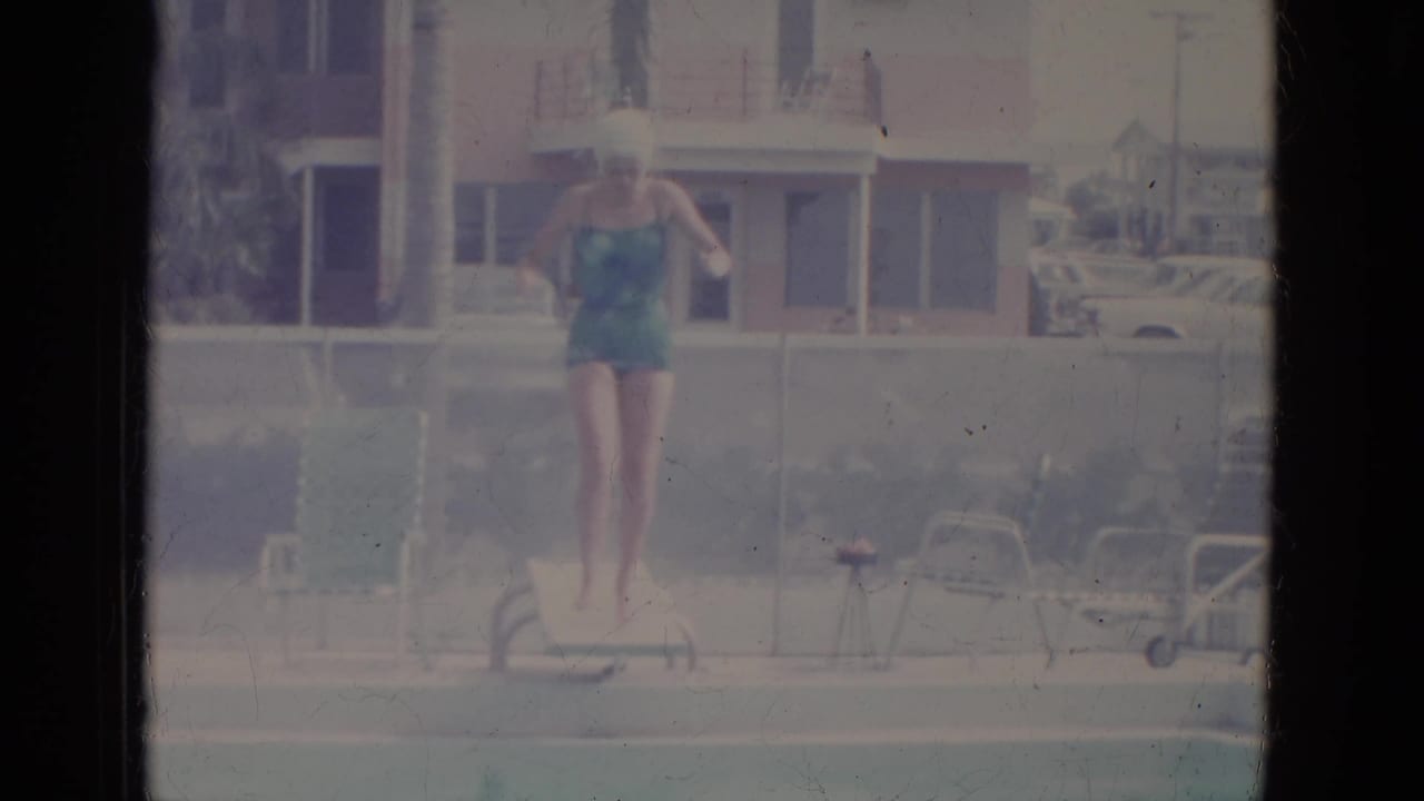 Woman rises from the diving board with pool furniture and a building in the background.