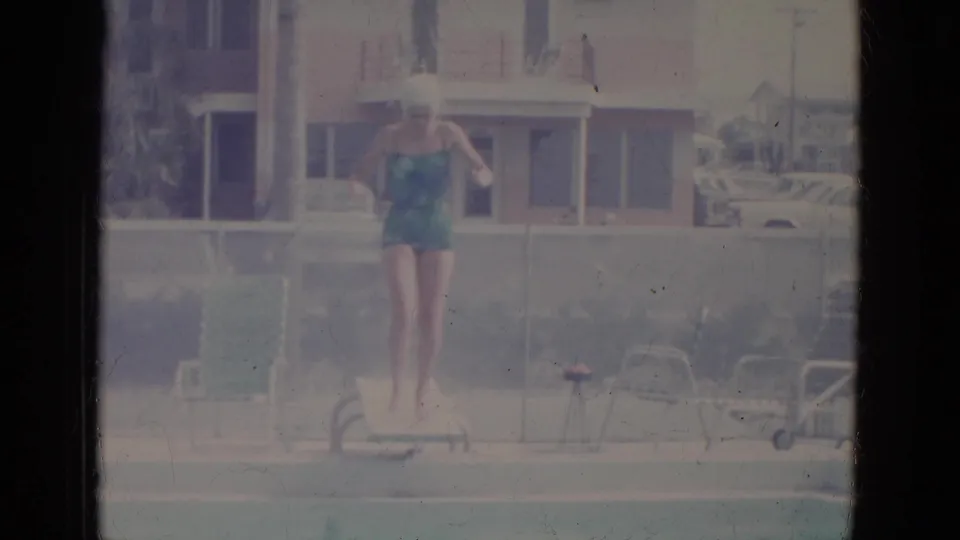 The woman in a teal swimsuit appears mid-jump from the pool diving board, with lounge furniture and a building visible behind her.