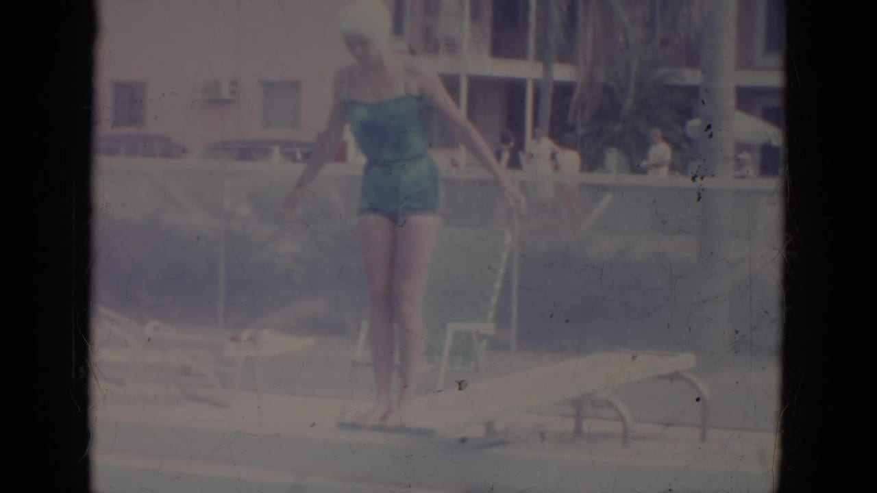 Woman in a teal swimsuit stands at a pool edge with arms raised near lounge chairs.