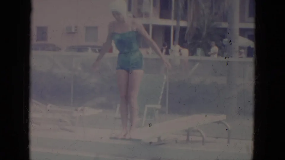 In supported 1966 Florida home-movie footage, a woman in a teal one-piece swimsuit and white cap prepares to dive at a pool edge.