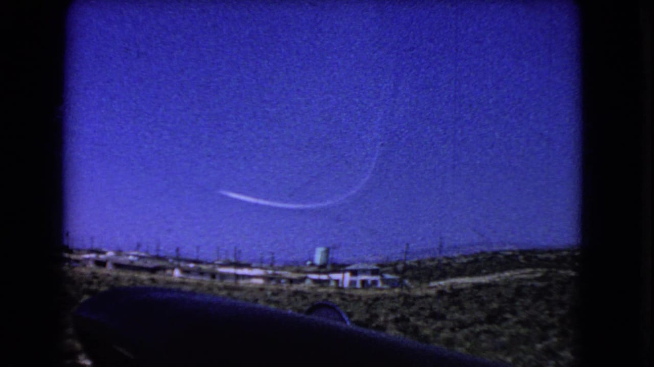1966 California 8mm frame showing pale contrails in a deep blue sky above arid hills, low buildings, and a green water tower.