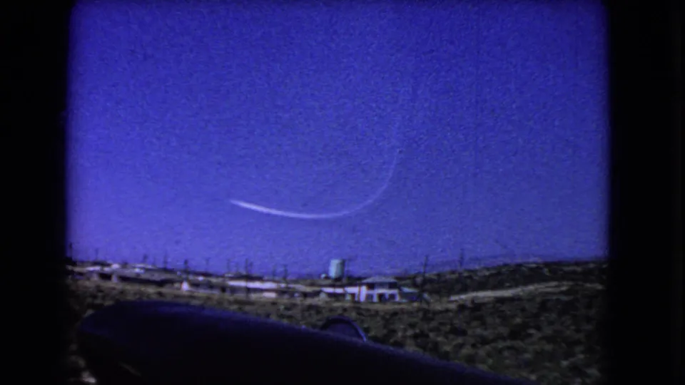 1966 California 8mm frame showing pale contrails in a deep blue sky above arid hills, low buildings, and a green water tower.