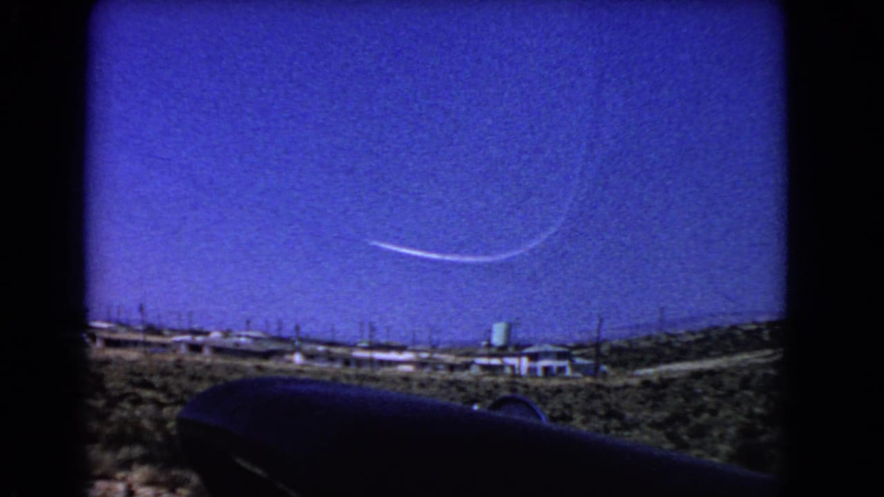 Curving contrail above desert hills, low buildings, and a water tower.