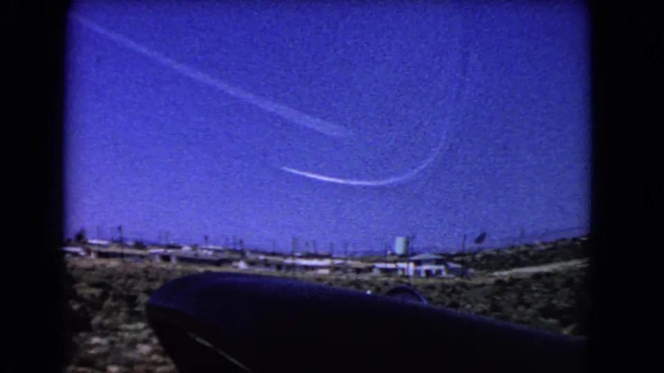 1966 California 8mm frame with two visible contrails crossing deep blue sky above arid ground, buildings, and a green water tower.