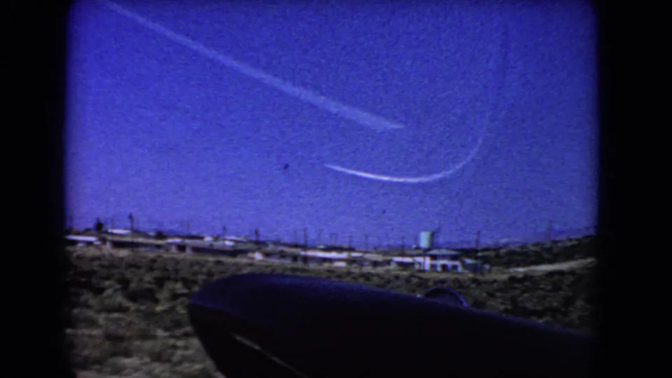 1966 California desert frame showing intersecting pale contrails over a small settlement, utility poles, and a green water tower.