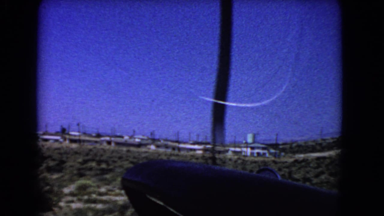 1966 California airplane-window view over arid terrain, with a curved contrail in deep blue sky and low structures on the horizon.