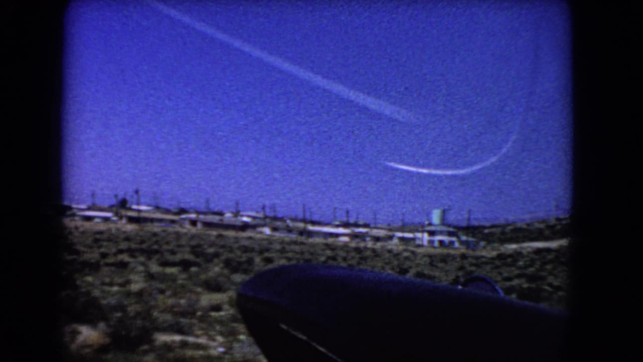 Desert landscape seen from an aircraft window, with bright sky, contrail, settlement, and dark foreground silhouette.