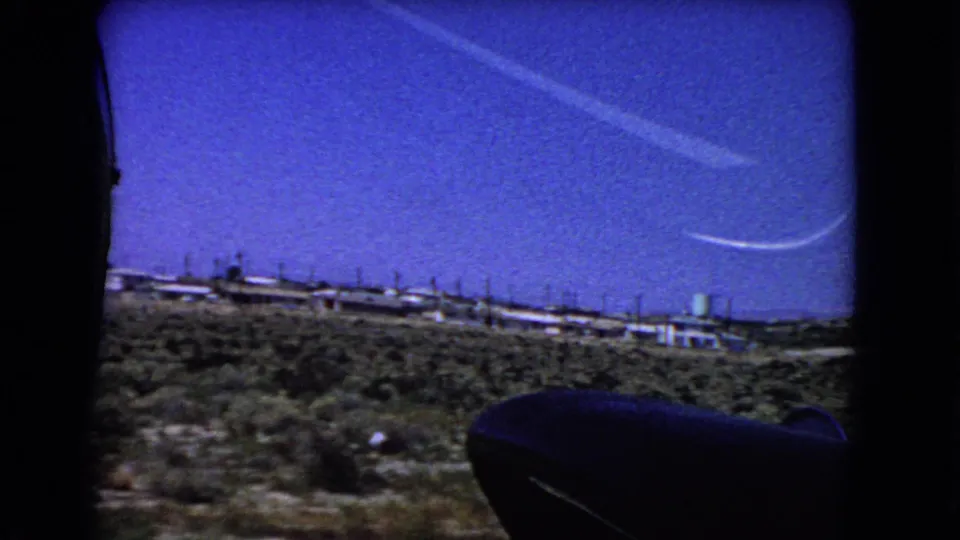1966 California desert landscape viewed from an airplane window, with low structures, scrubland, and a white contrail against saturated blue sky.