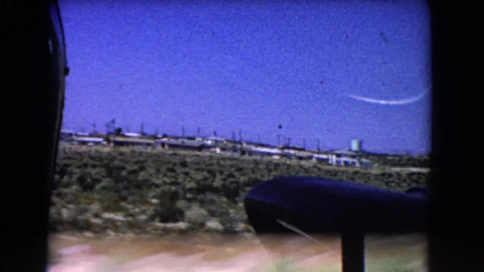 Final 1966 California airplane-window view with open desert below, low settlement on the horizon, and a faint curved contrail in the sky.