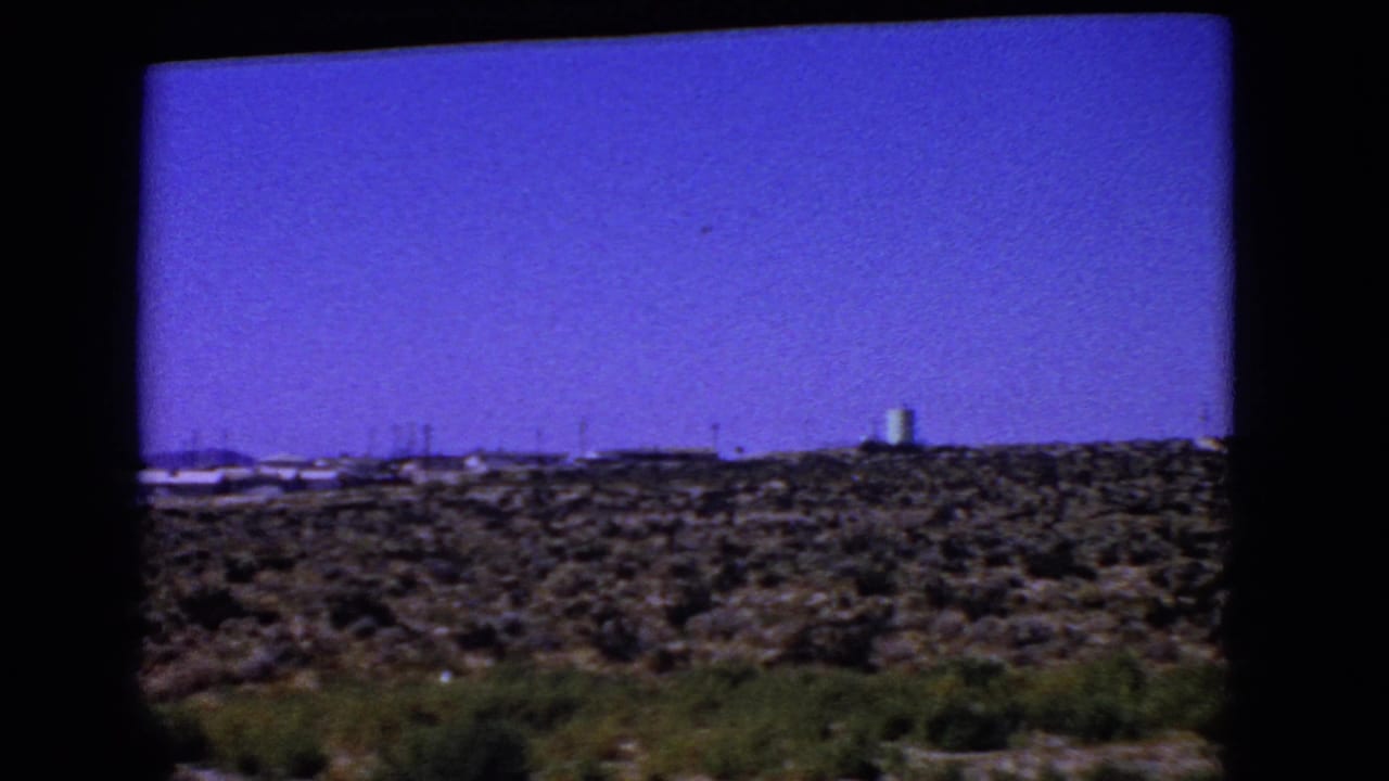 1966 California 8mm car-window view of scrubby desert terrain with distant industrial structures on the horizon.