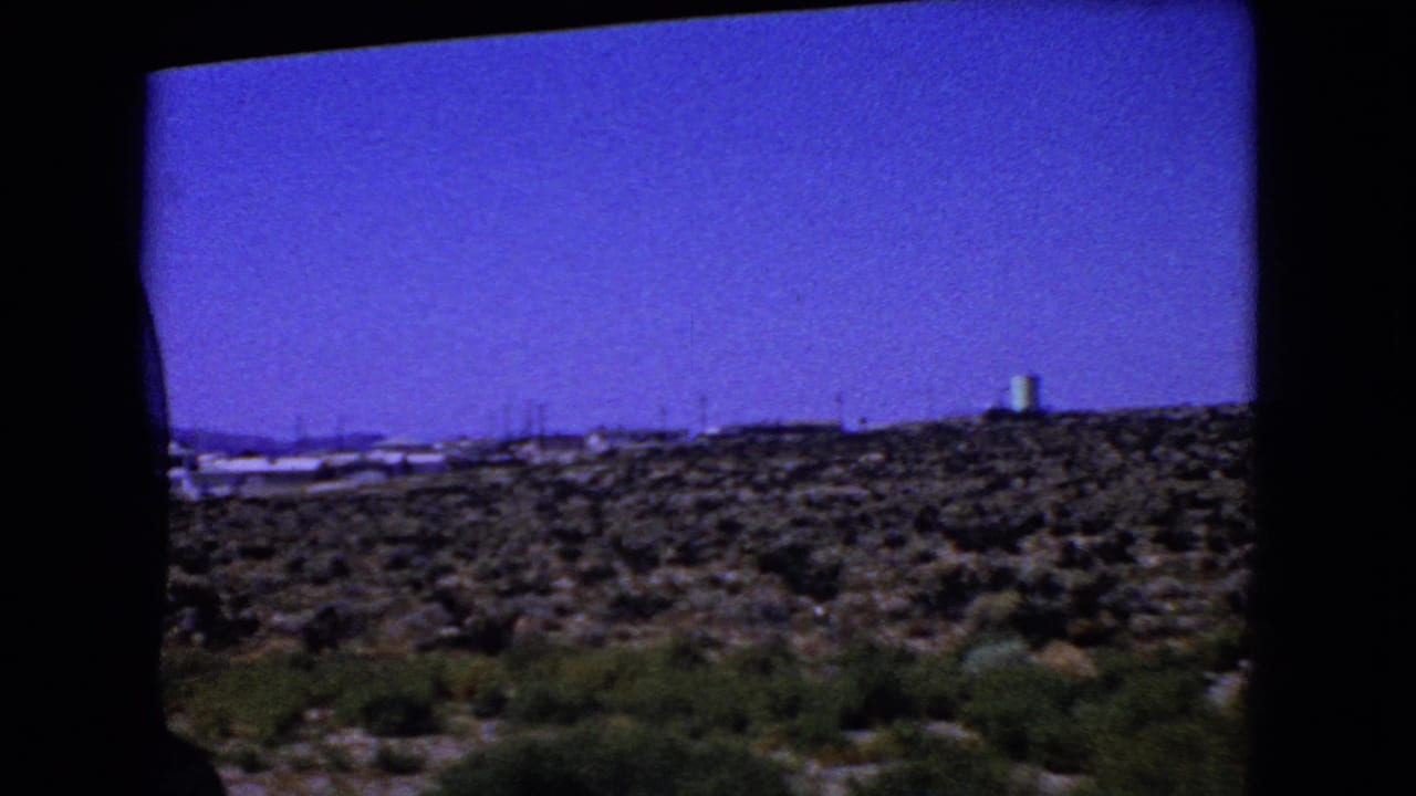 1966 California home-movie frame looking past a dark car-window edge to desert scrub and a distant pale tower.