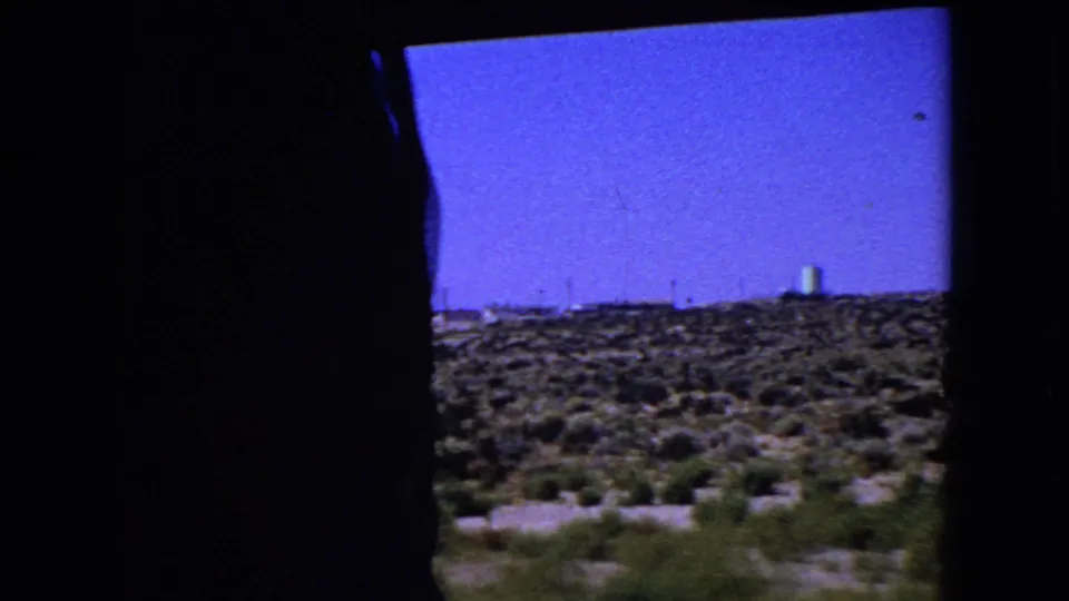 1966 California 8mm frame showing desert scrub, a distant industrial horizon, and dark vehicle framing at the edge.