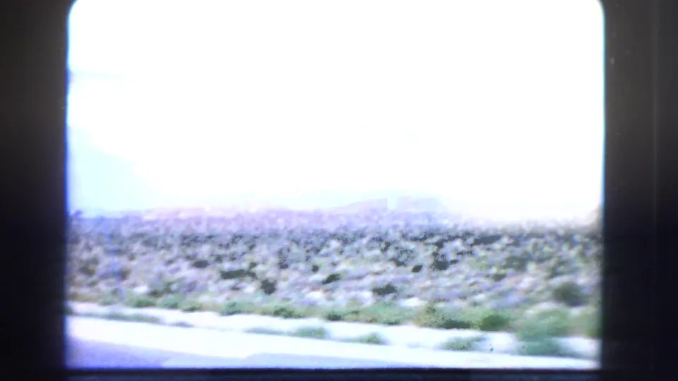 Brightly exposed 1966 California 8mm frame with desert brush, road edge, and distant horizon structures through a car window.