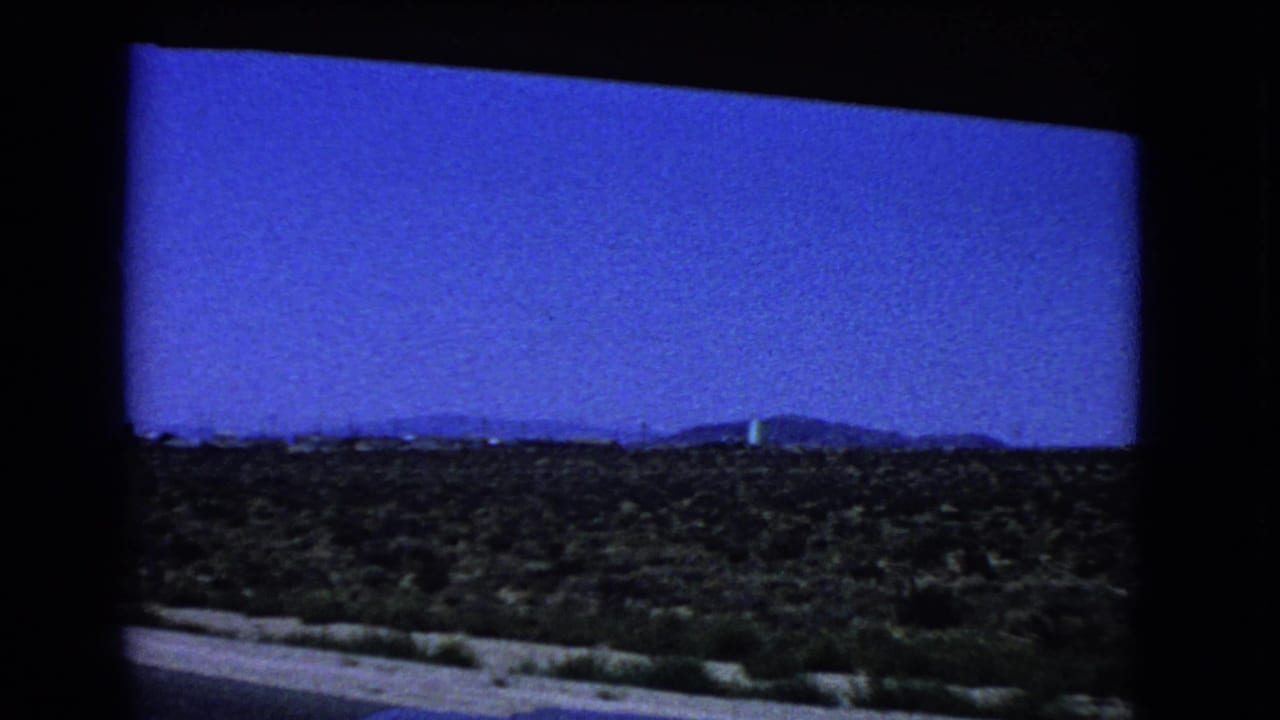 Desert scrubland seen past a road edge with distant hills under a blue sky.