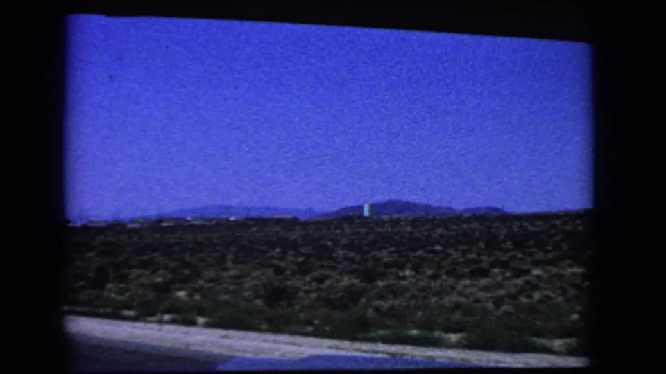1966 California 8mm frame of a roadside desert landscape, with scrub vegetation, low hills, and saturated blue sky.