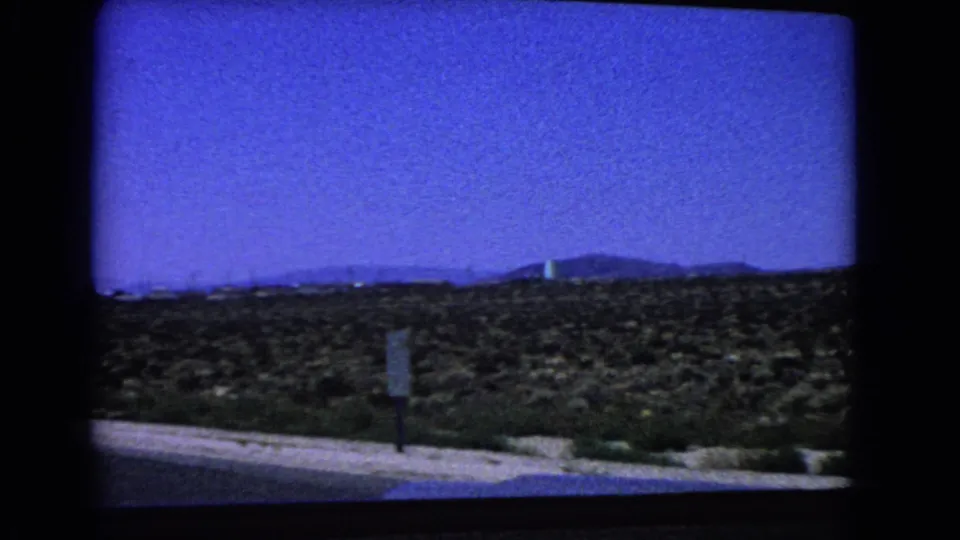 1966 California 8mm frame with a small roadside sign in the foreground, desert scrub beyond, and low hills under a blue sky.