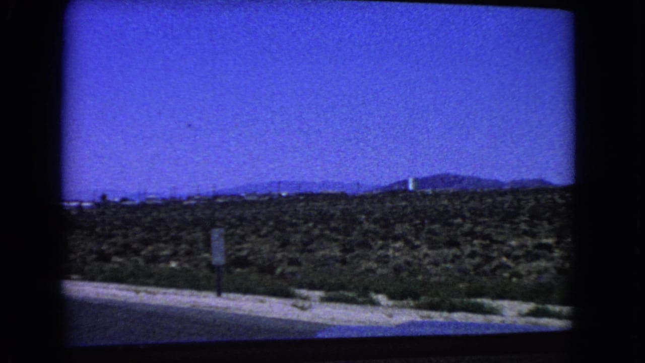 1966 California desert roadside seen from a moving car, with scrubland, a small road sign, distant mountains, and dark film-frame edges.