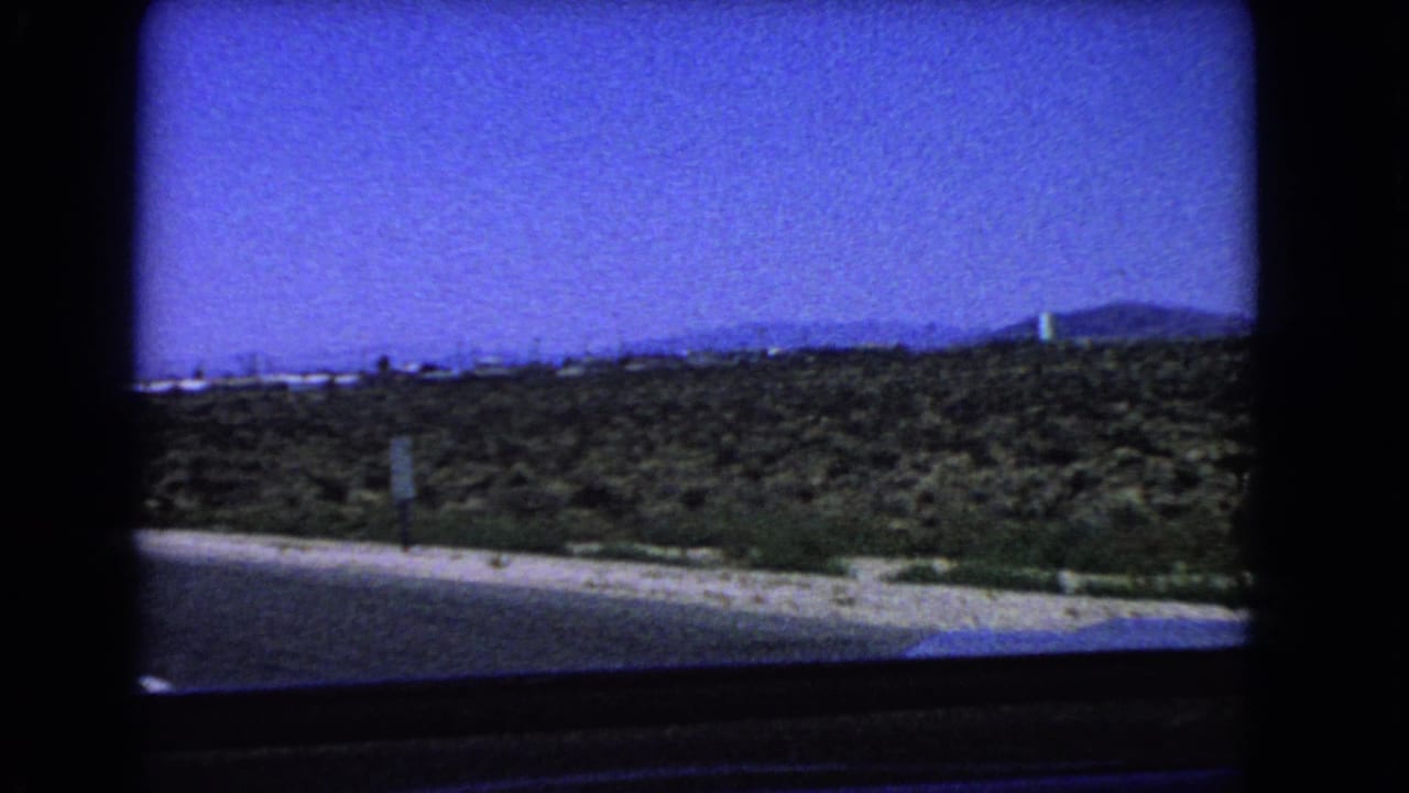 Road shoulder and desert scrub seen from a moving car, with mountains on the horizon.