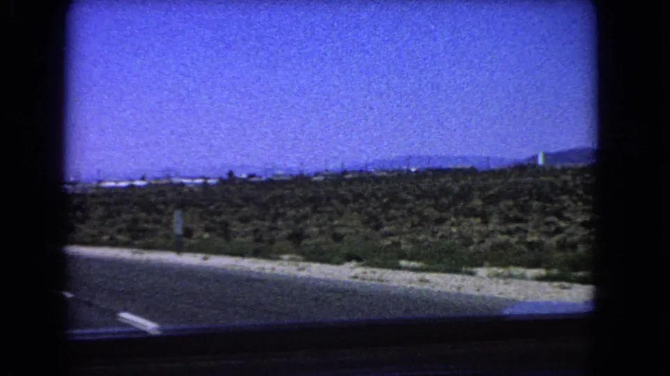 California desert roadside in 1966 with scrubland, a small sign, low hills, a distant white tower, and visible 8mm frame borders.