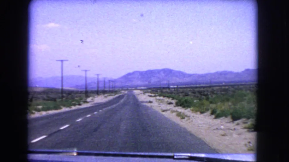1966 California desert road seen from a moving car, with lane markings, utility poles, scrub shoulders, mountains, and the car hood at the bottom.