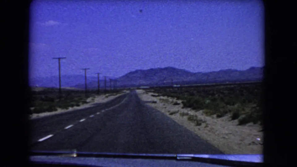 1966 California desert road viewed from inside a moving car, with utility poles along the highway and mountains ahead.