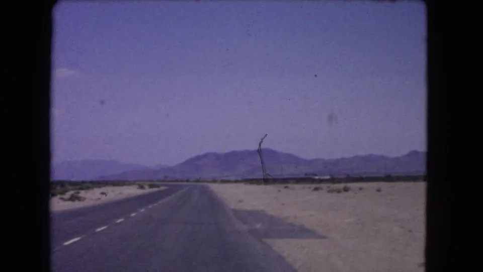 1966 California desert driving view with a two-lane road, open sand, distant mountains, and a leaning roadside pole.