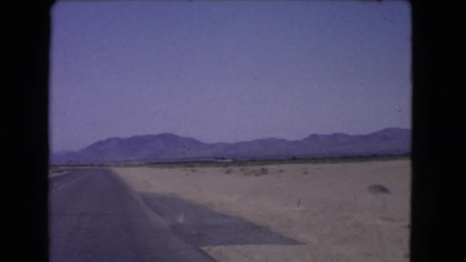 Faded 8mm view of a California desert road in 1966, showing a broad shoulder, scrubland, and mountains across the horizon.