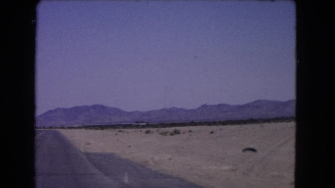 1966 8mm view from a moving car across a California desert highway edge toward scrubland and distant mountains.