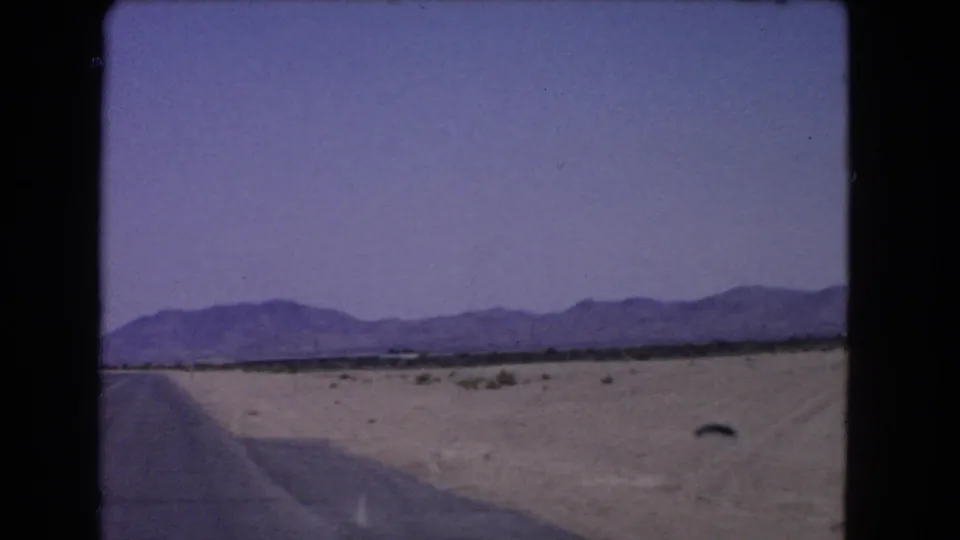 1966 8mm view from a moving car across a California desert highway edge toward scrubland and distant mountains.