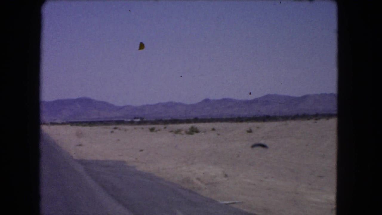 Passenger-window view of a California desert road with sparse vegetation and mountains on the horizon in 1966 8mm footage.