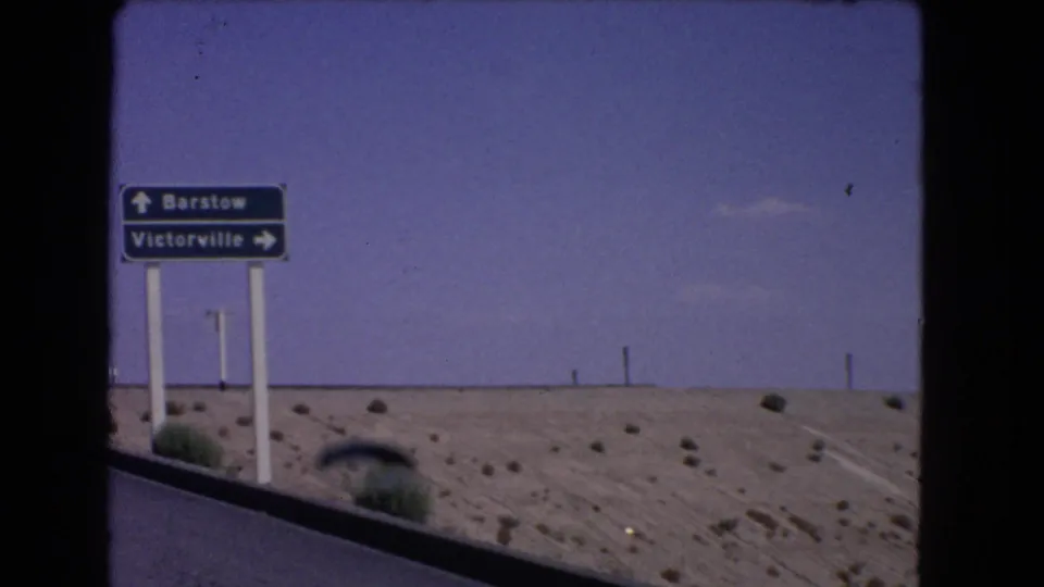 1966 California desert roadside sign points toward Barstow and Victorville beside sand, scrub, utility poles, and blue sky.