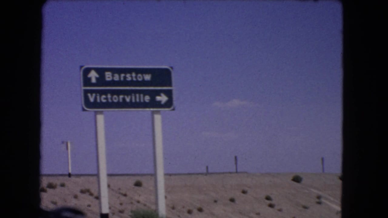 1966 California home-movie frame showing a directional sign for Barstow and Victorville against blue sky and arid roadside terrain.