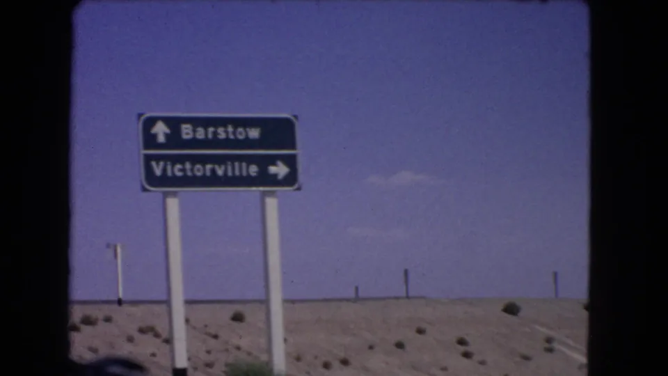 1966 California home-movie frame showing a directional sign for Barstow and Victorville against blue sky and arid roadside terrain.
