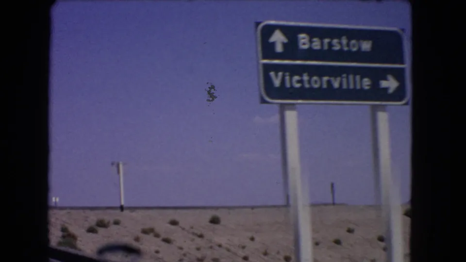 Closer moving-vehicle view of a California directional road sign for Barstow and Victorville, with desert scrub and sky behind it.