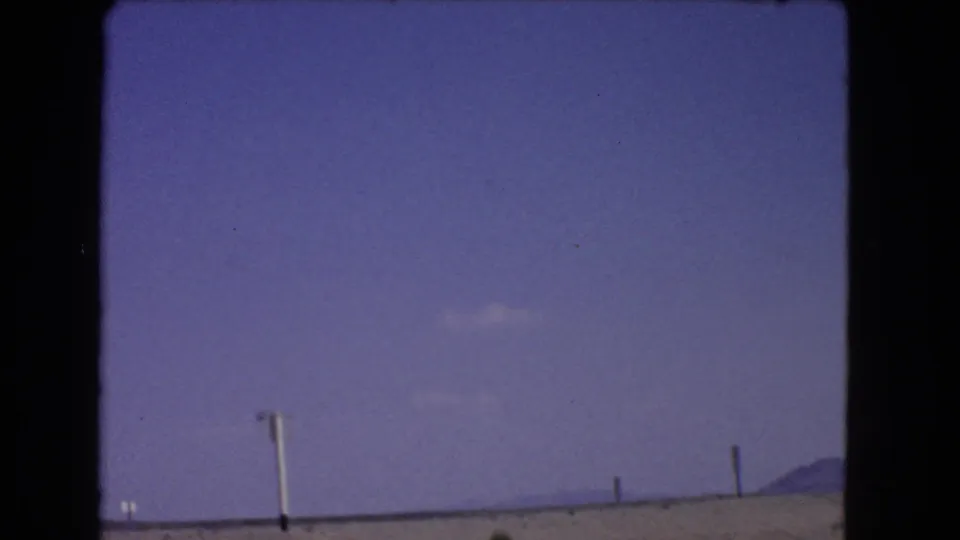 After passing the sign, the frame shows open blue sky, arid California roadside terrain, sparse shrubs, and small roadside posts.
