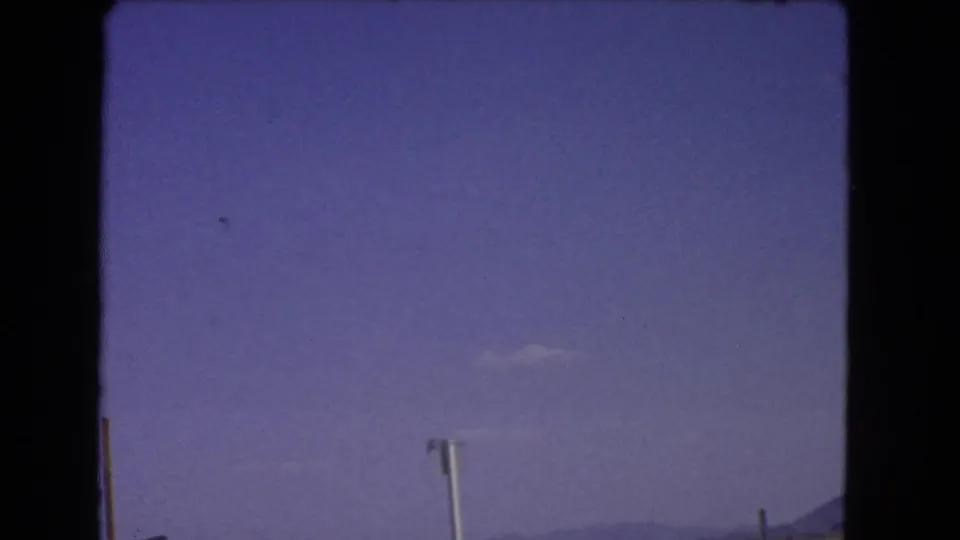 Final 1966 California roadside frame with open sky, dry desert ground, a partial signpost or roadside marker, and distant low hills.