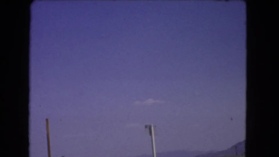 1966 California 8mm frame showing an arid roadside edge, upright poles, hazy distant mountains, and a blue-purple sky from a moving vehicle.
