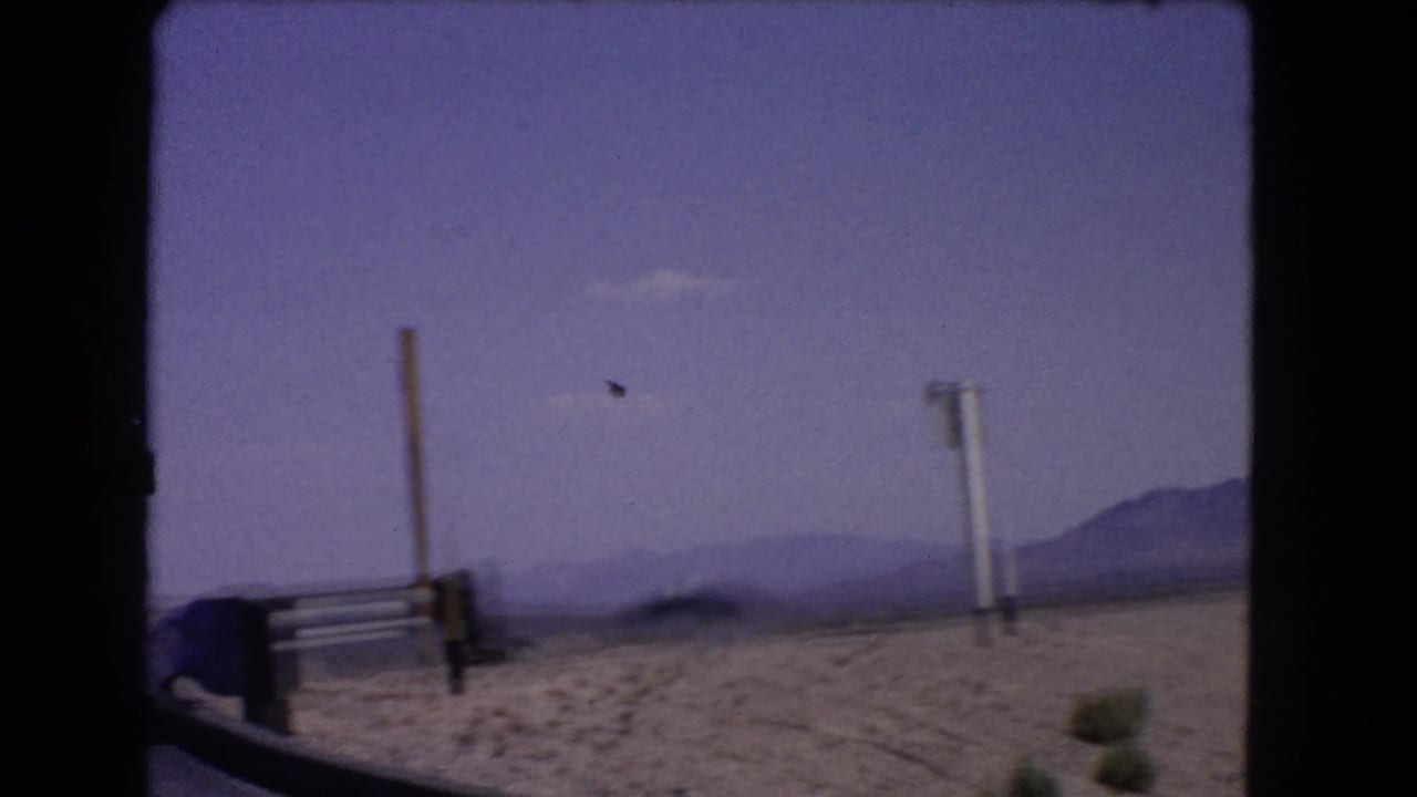1966 California roadside view with sandy desert ground, upright posts, distant mountain ridges, and a small dark object crossing the sky.