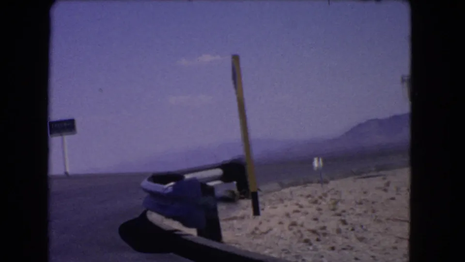 1966 California 8mm view from a moving vehicle, showing a highway edge, small roadside sign, pole, dry ground, and distant mountains.