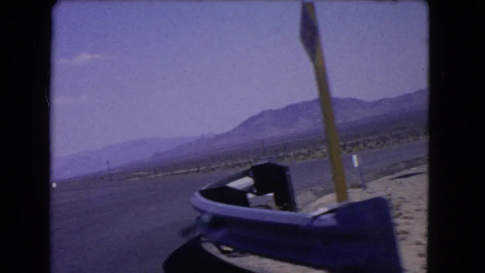 1966 California highway-side frame with a close pole, paved road edge, dark vehicle frame, dry roadside terrain, and distant mountains.