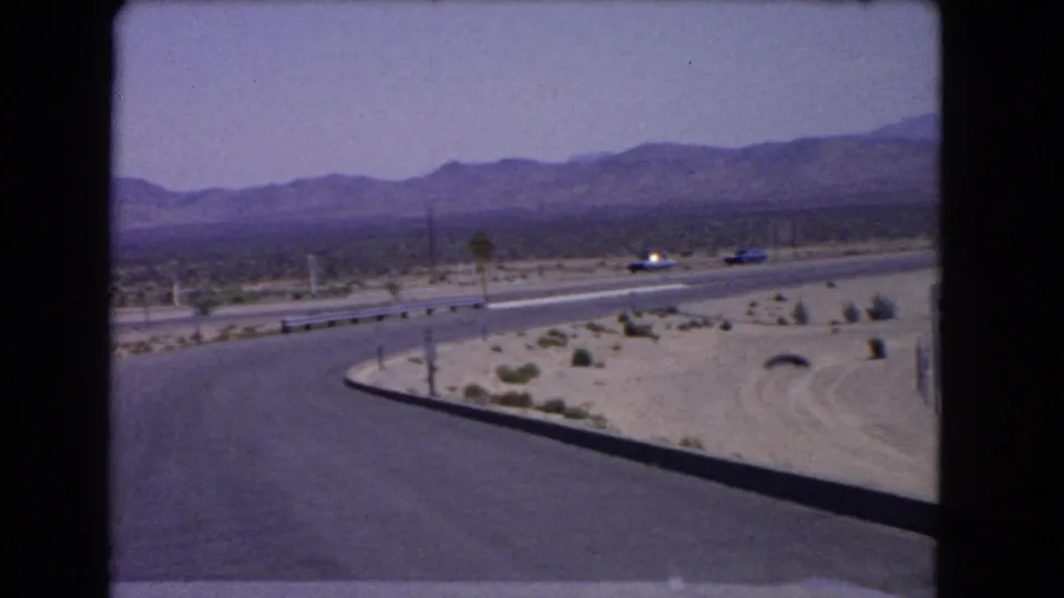 Wide 1966 California home-movie view of a desert road, highway traffic, sparse planting, and mountain backdrop.