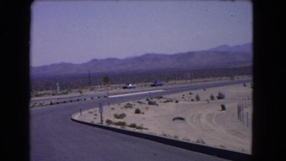 1966 California 8mm frame showing classic cars traveling across an arid highway with mountains behind.