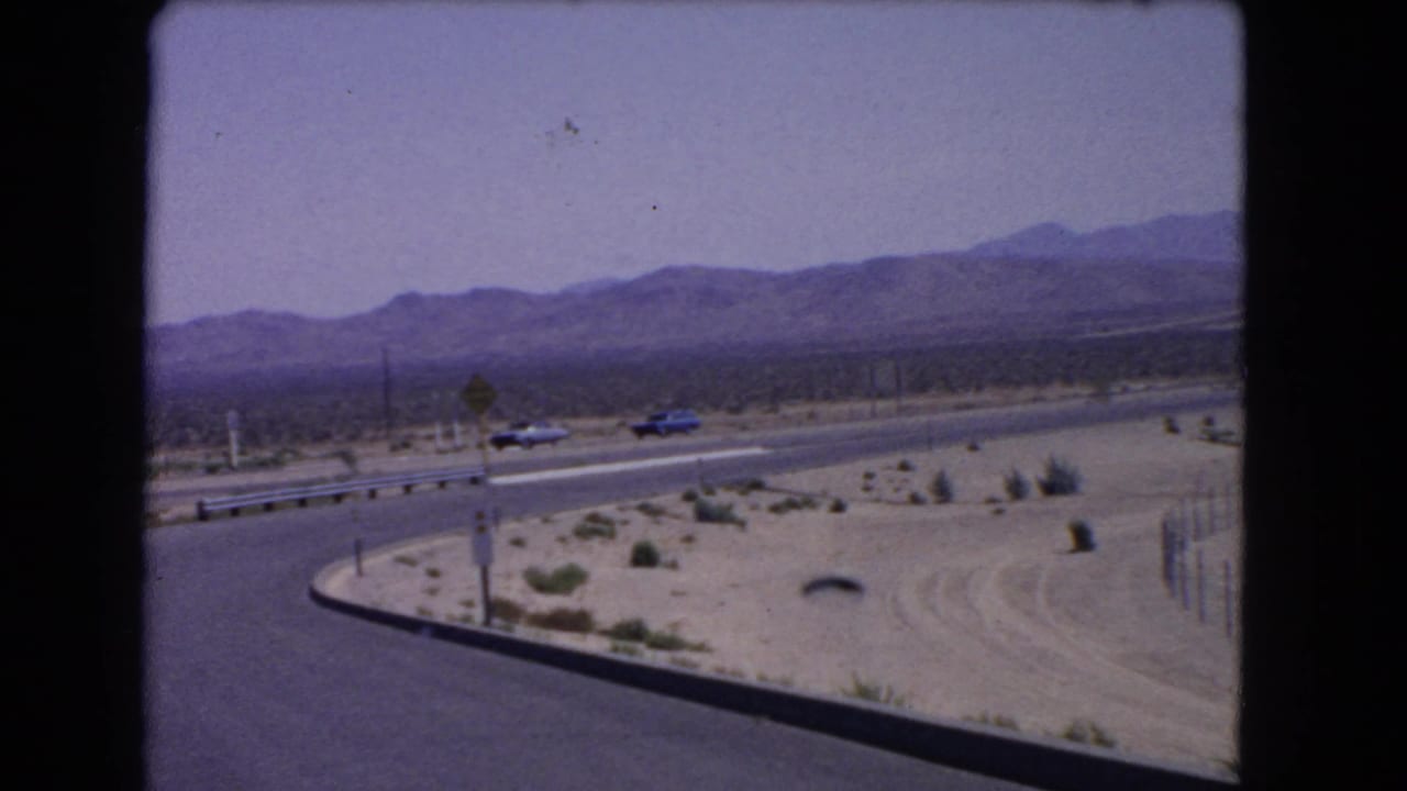 1966 California 8mm frame of cars on a curved desert highway with sandy terrain, sparse shrubs, roadside signs, and mountains in haze.