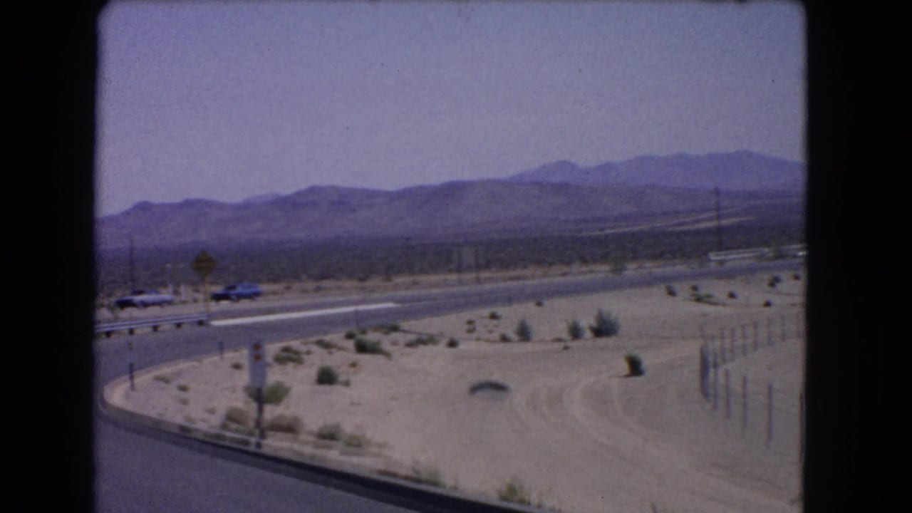 Desert roadside and curved highway with cars, warning sign, guardrail, shrubs, and mountains.