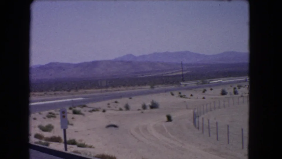 1966 California 8mm frame with a roadside sign in the foreground, a curving highway, desert fencing, shrubs, and hazy mountain backdrop.