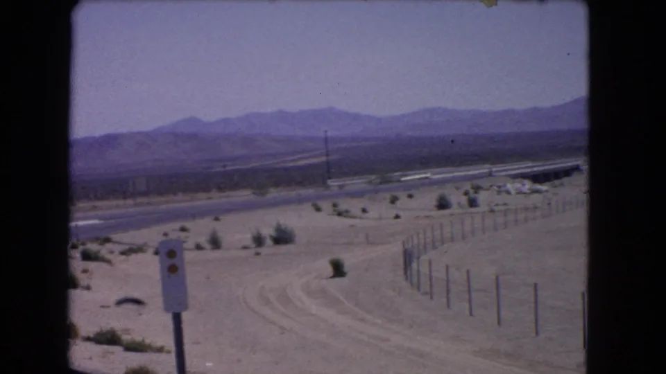 1966 California desert highway view with a foreground sign, fence line, sandy shoulder, low shrubs, guardrail, and mountains under a hazy sky.