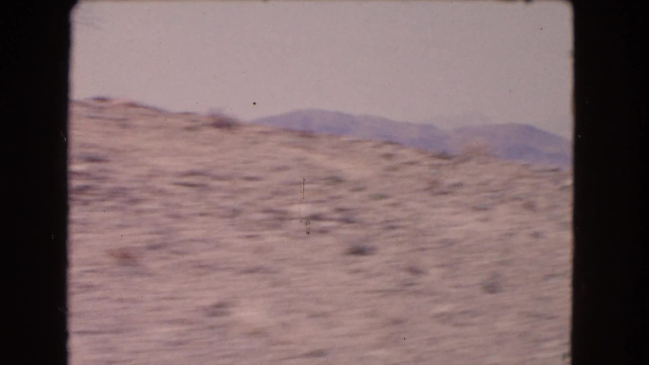 Blurred desert hillside and distant mountains seen through a moving vehicle window.