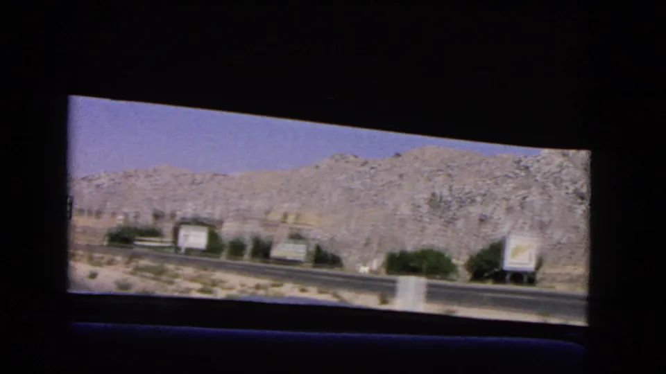 1966 California roadside scene with billboards, a guardrail, sparse shrubs, paved road, and rocky desert hillside.